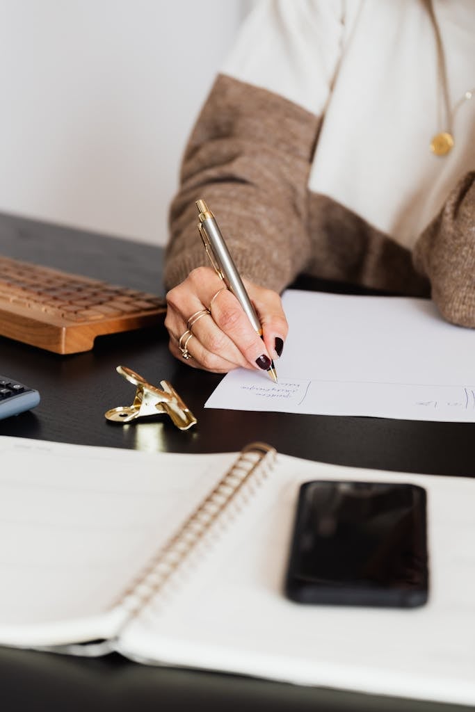 Unrecognizable female entrepreneur writing on piece of paper while sitting at table with notebook and smartphone