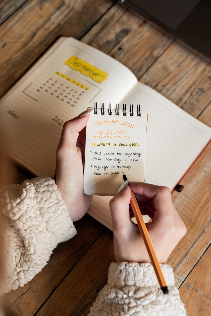 A person holds a notepad jotting down a to-do list with a calendar in the background.