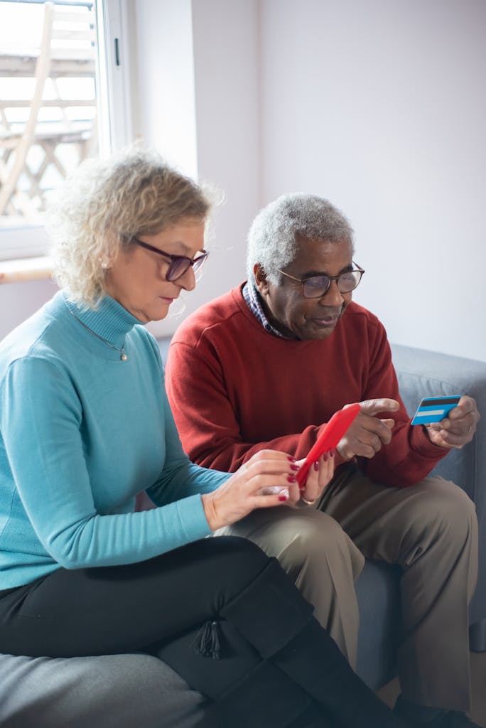 Senior couple sitting indoors, using smartphones and credit cards for online shopping.