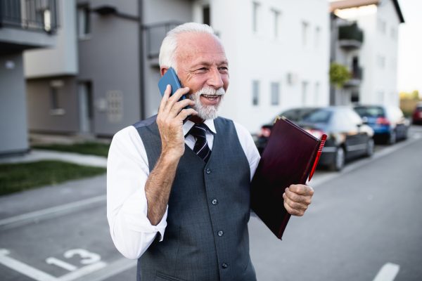 Real estate agent standing in front of a new house for sale and talking on his smart phone.